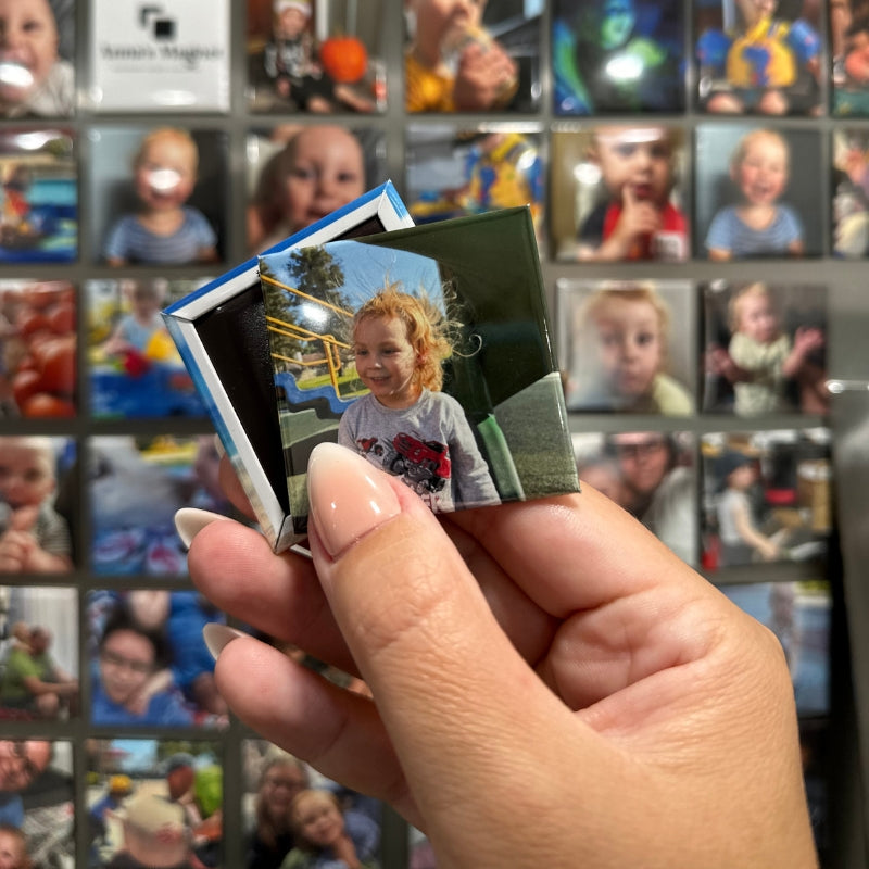 Hand holding 2x2 custom photo magnet of a child in front of a grid of other custom phot magnets