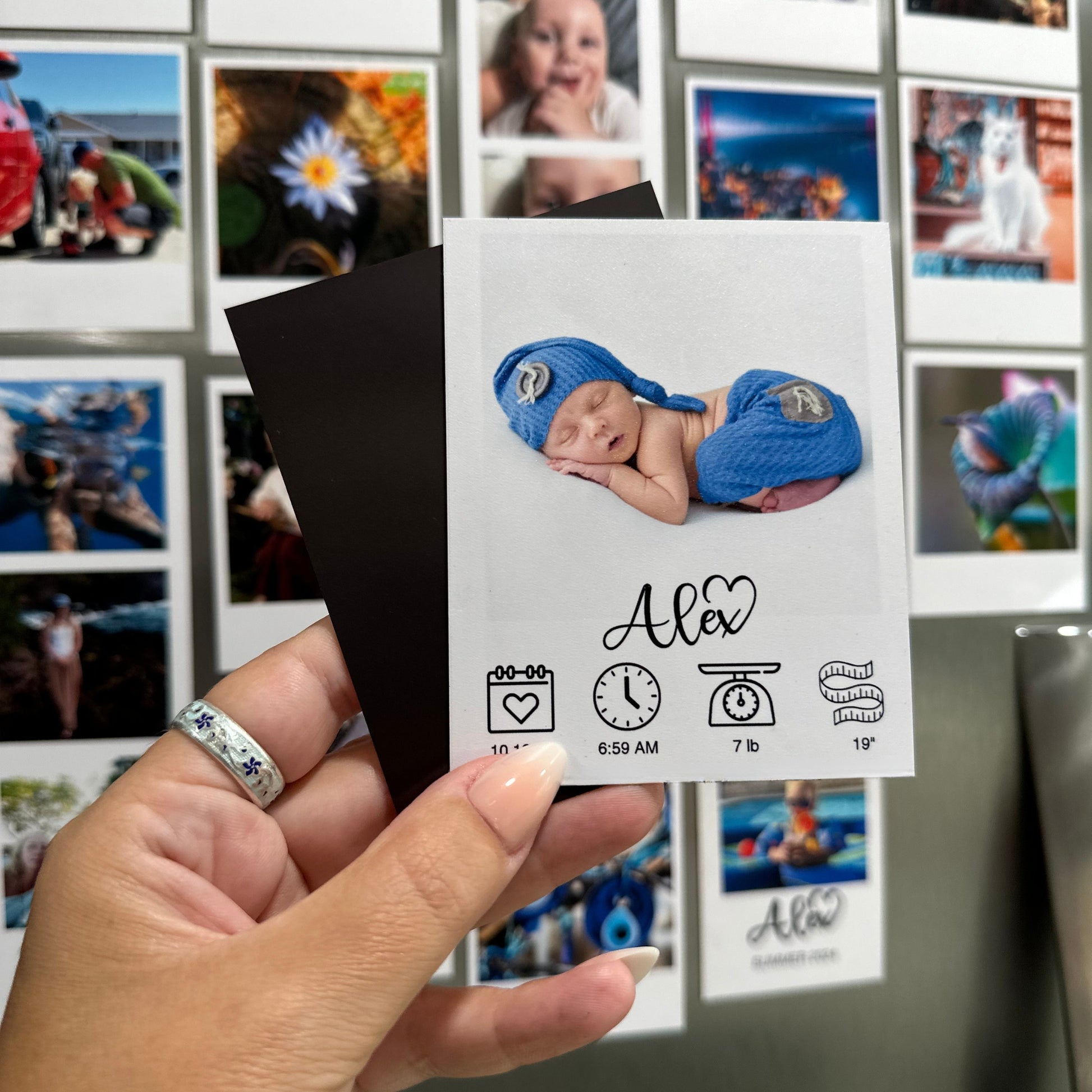 Hand holding a baby milestone custom photo magnet with a photo of a baby in blue booties against a stainless steel refrigerator with other custom photo magnets.