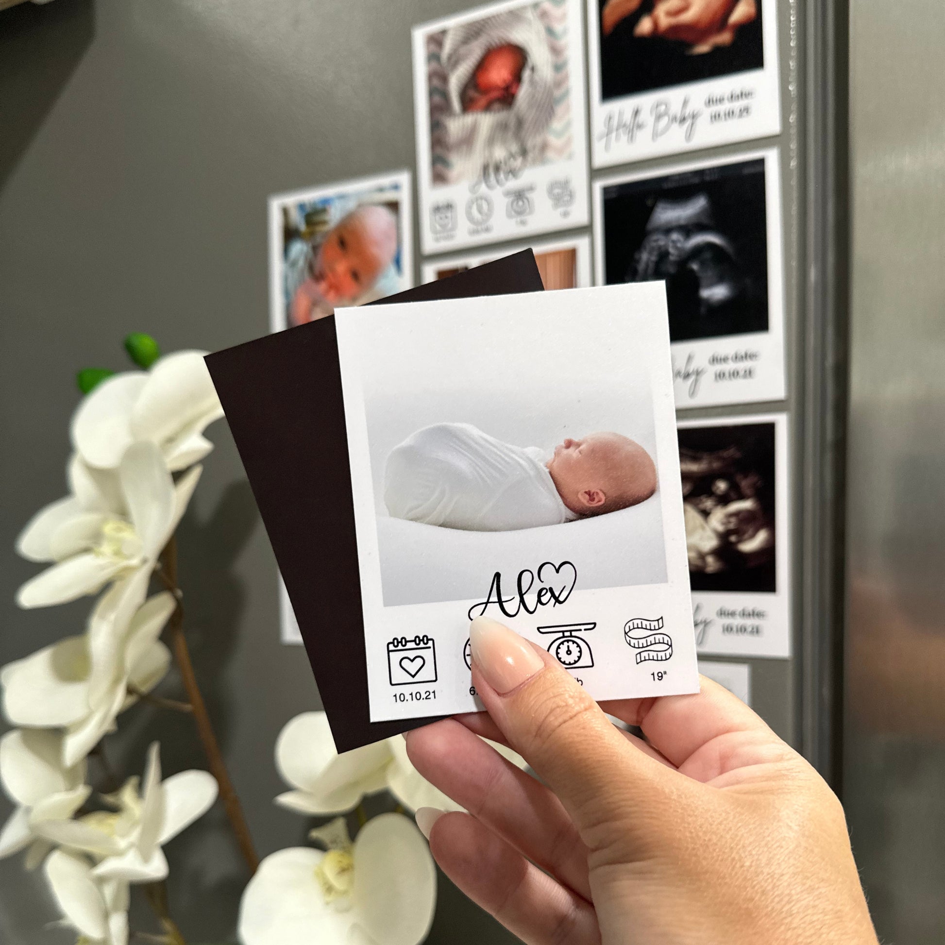 Hand holding a baby milestone custom photo magnet with a photo of a baby in white swaddle against a stainless steel refrigerator with other custom photo magnets.