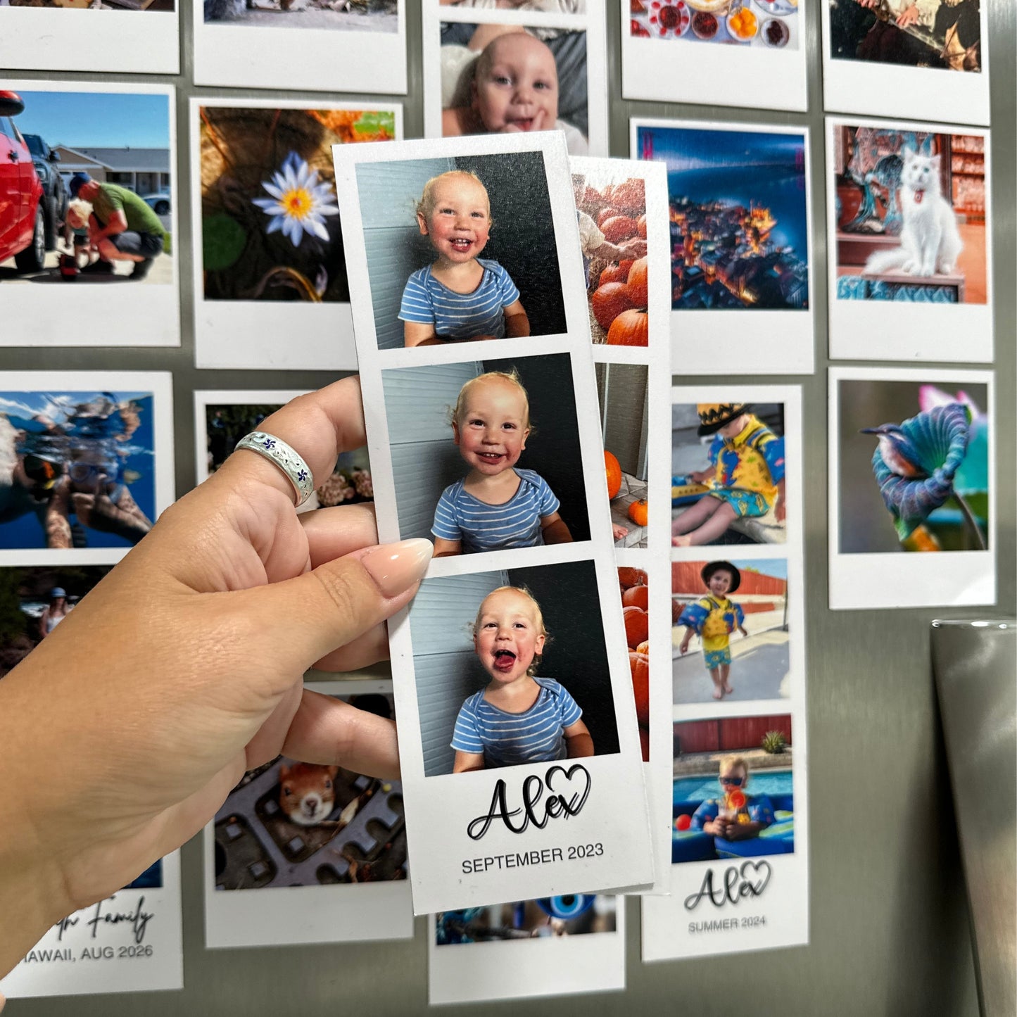 Hand holding a custom photo booth strip magnets with baby photos against a stainless steel refrigerator with other custom photo magnets.