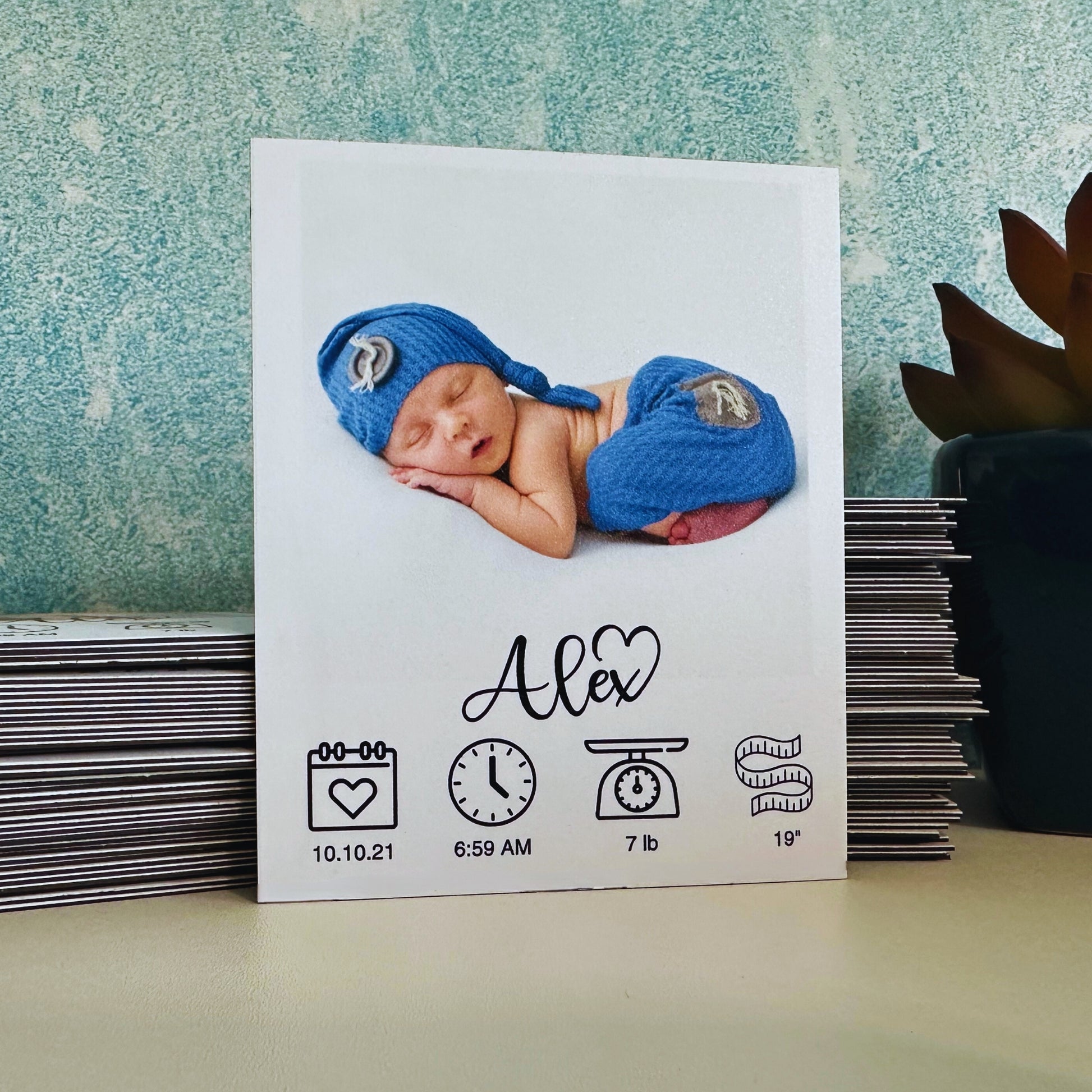 Newborn baby in blue outfit with birth details on a custom photo magnet against a textured wall.