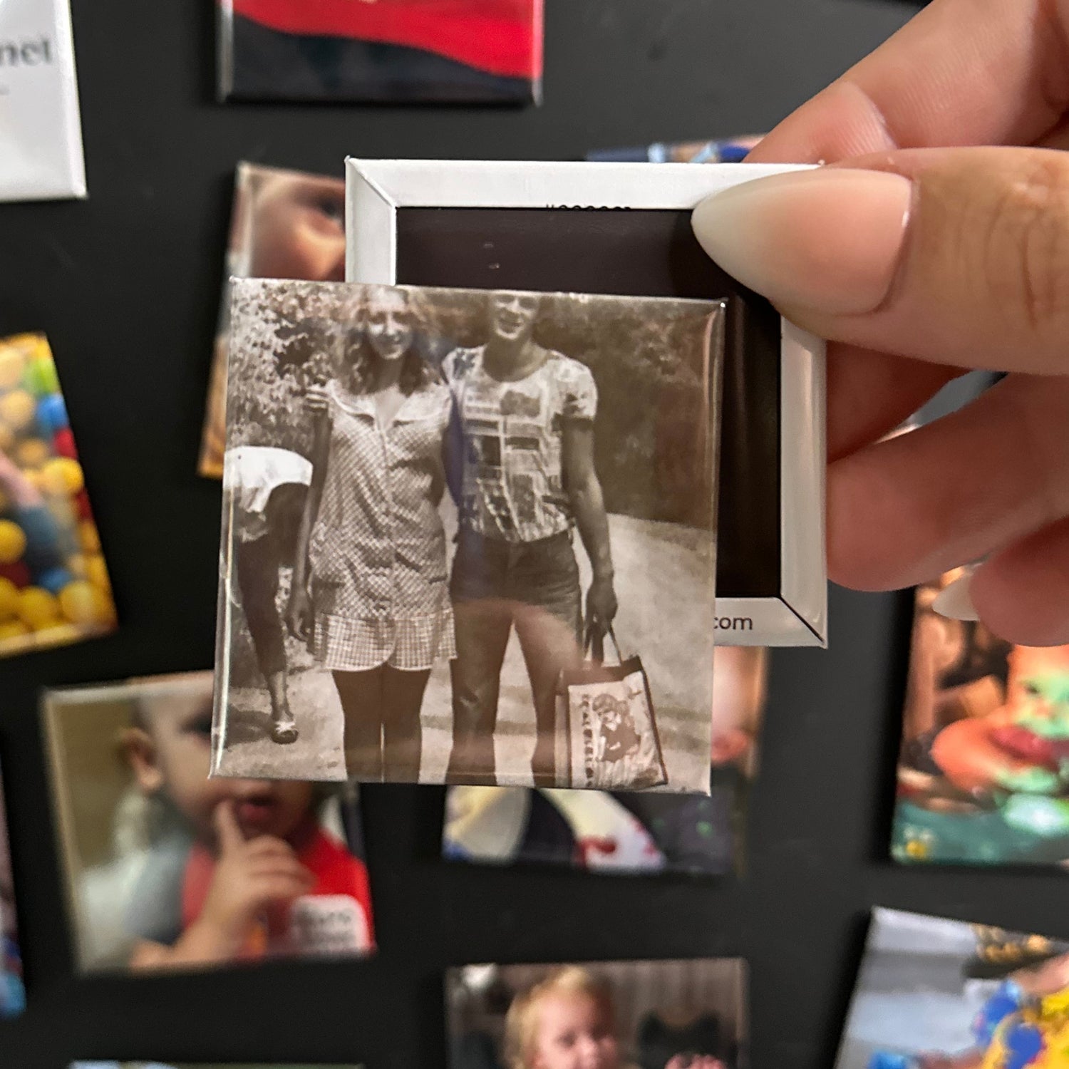 Person holding a custom 2x2 photo magnet with black and white photo of two people in front of a collage of other custom photo magnets.