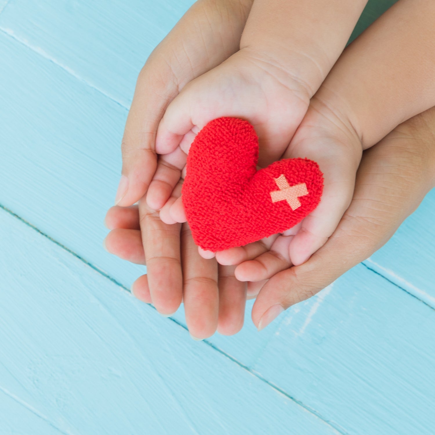 Child's hands holding a red heart with a bandage on a light blue wooden surface