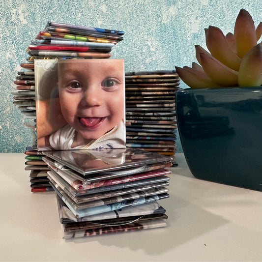 Stack of 2x2 custom photo magnets with a magnet with a child, placed on a surface with a plant in the background.