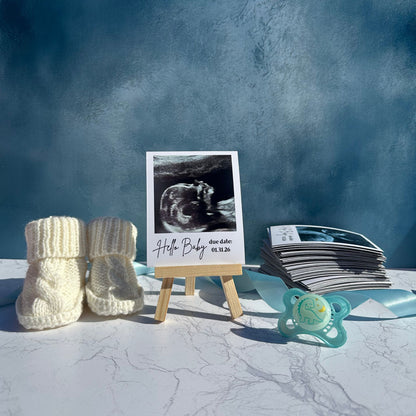 Baby booties, ultrasound photo magnet on a wooden stand, and pacifier with a blue background