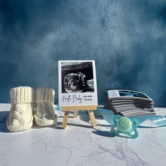 Baby booties, ultrasound photo magnet on a wooden stand, and pacifier with a blue background
