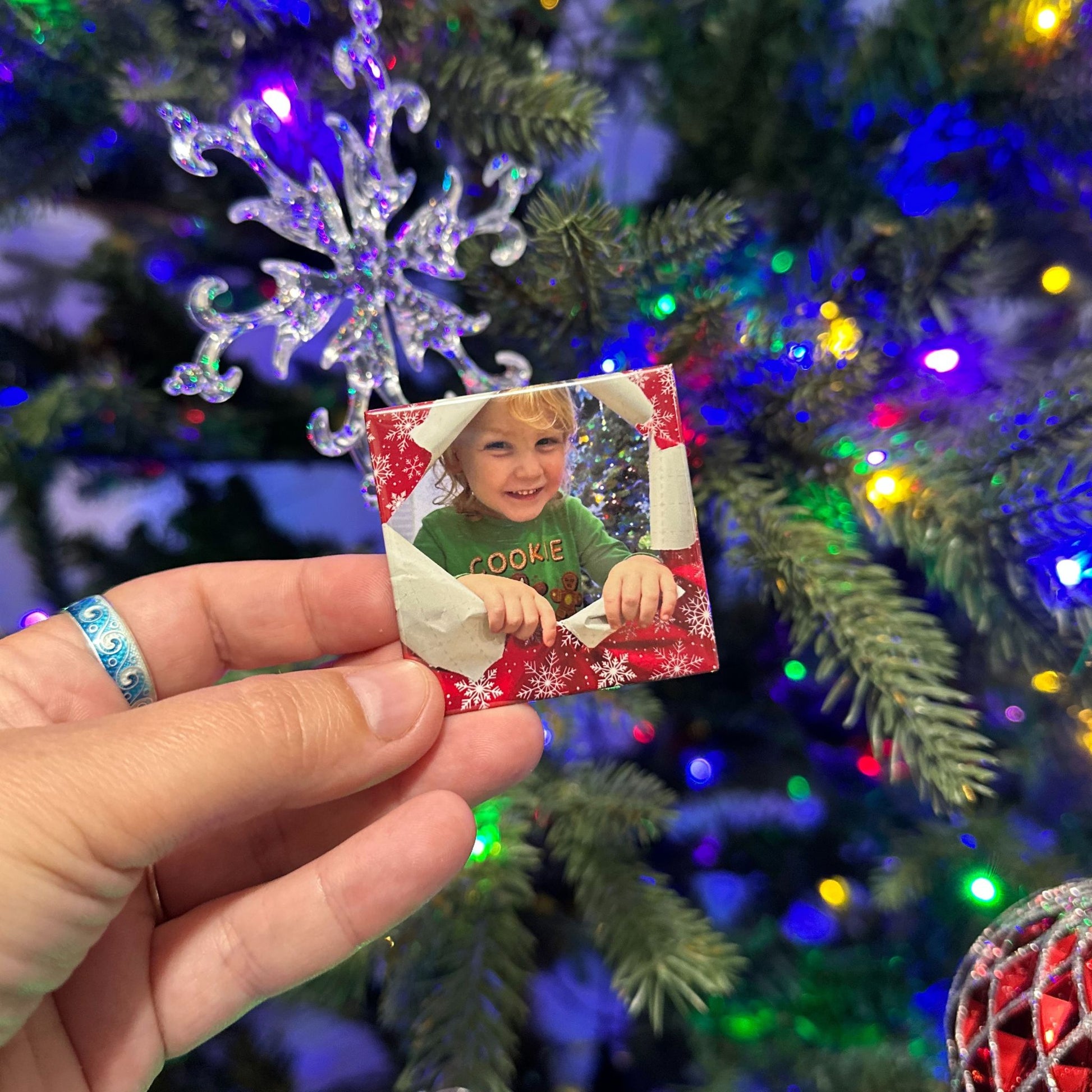 Hand holding a custom photo magnet with a child's picture in front of a decorated Christmas tree.