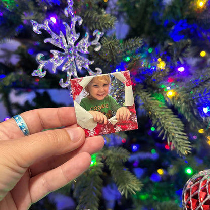 Hand holding a custom photo magnet with a child's picture in front of a decorated Christmas tree.