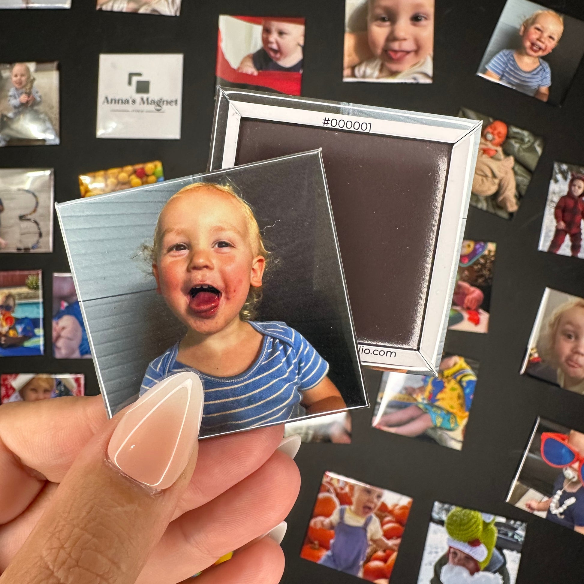 Hand holding a 2x2 custom photo magnet of a child with a tongue with a collection of other custom photo magnets in the background.