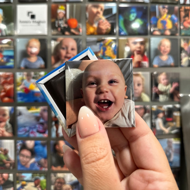 Hand holding a 2x2 custom photo magnet of a smiling baby in front of a grid of other custom photo magnets