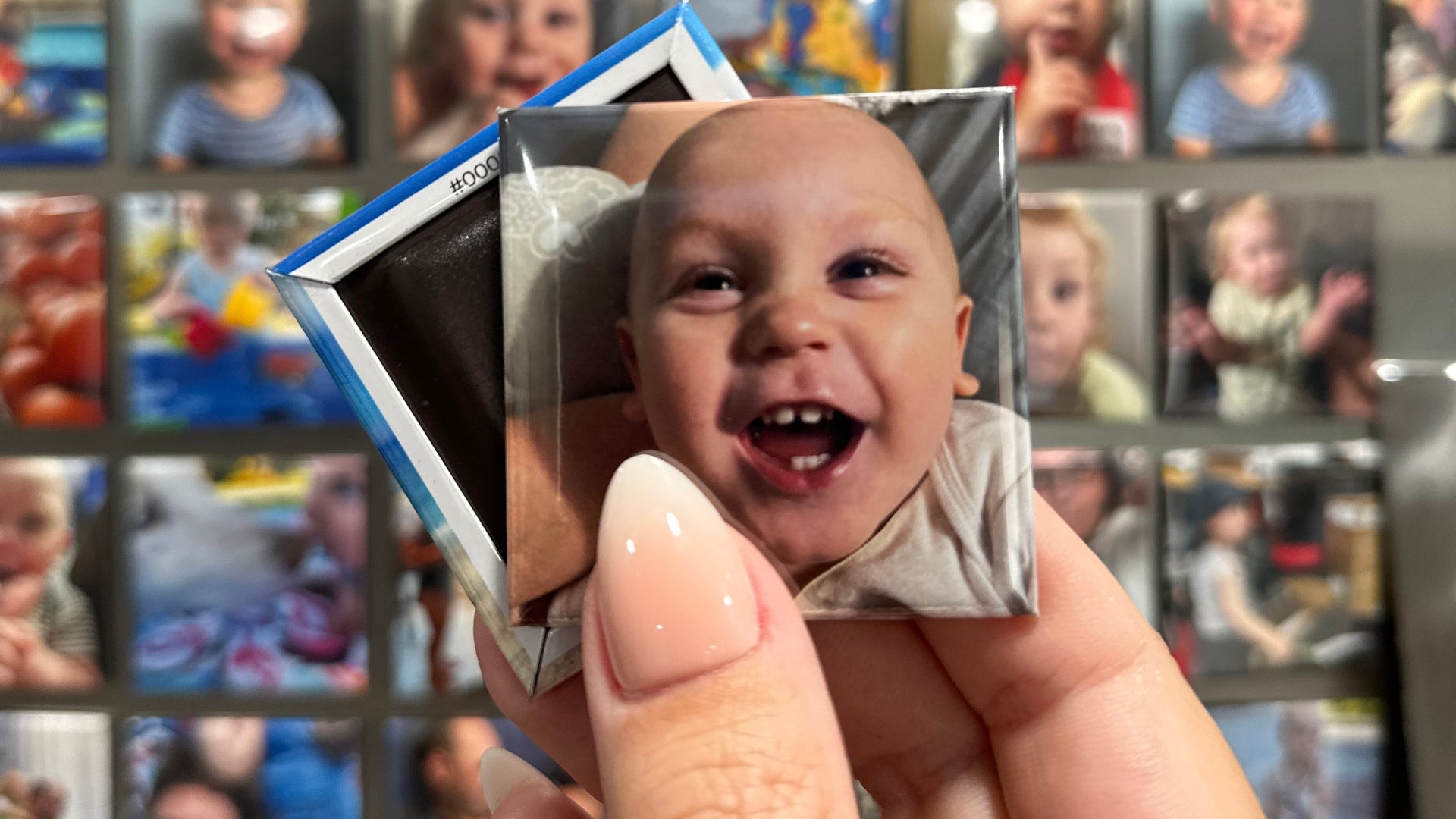 Hand holding a custom photo magnet of a baby with a grid of other custom made photos magnets  in the background