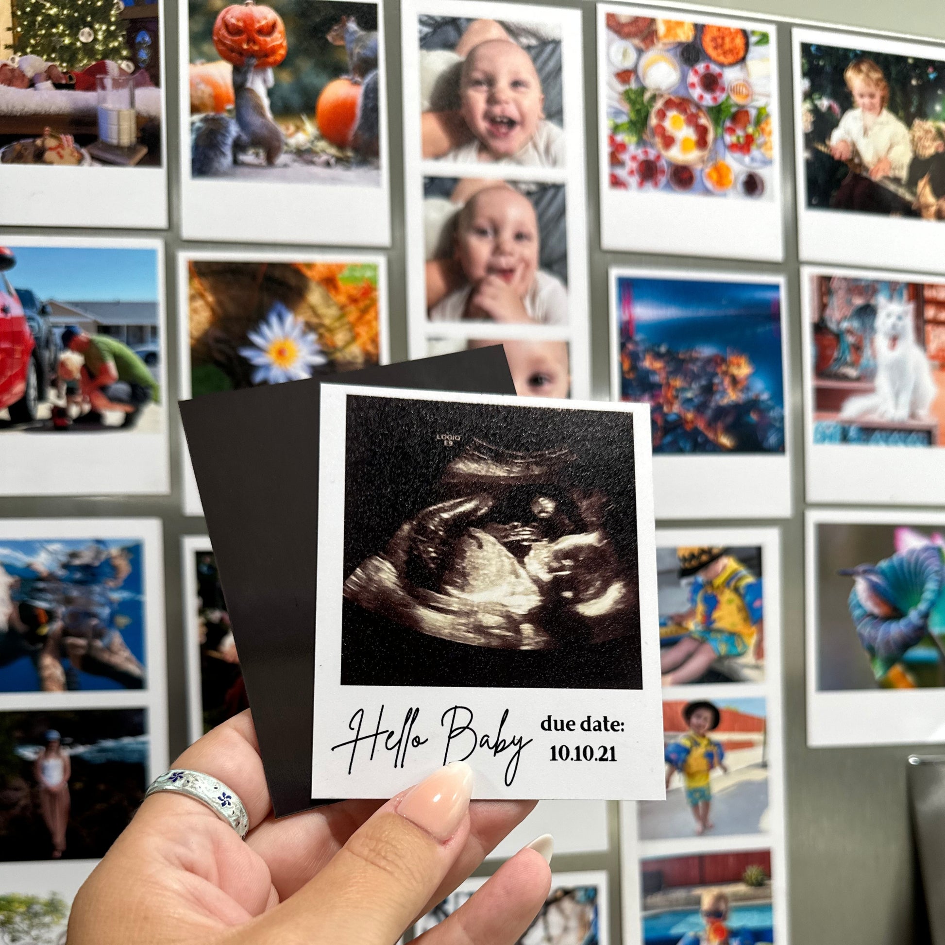Hand holding a custom photo magnet of an ultrasound with 'Hello Baby' text against other custom photo magnets on a refrigerator.