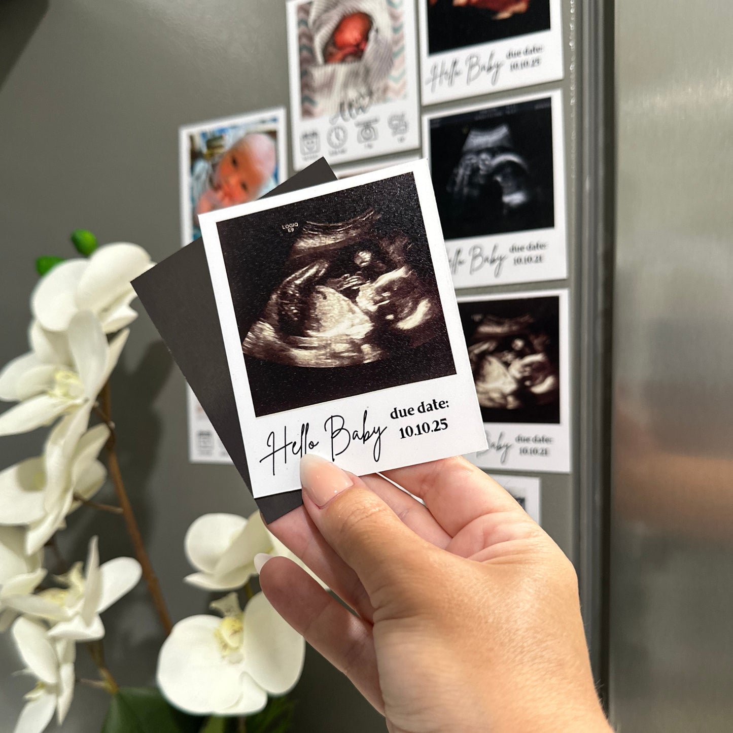 Hand holding a custom photo magnet of an ultrasound with 'Hello Baby' text against other custom photo magnets on a refrigerator.