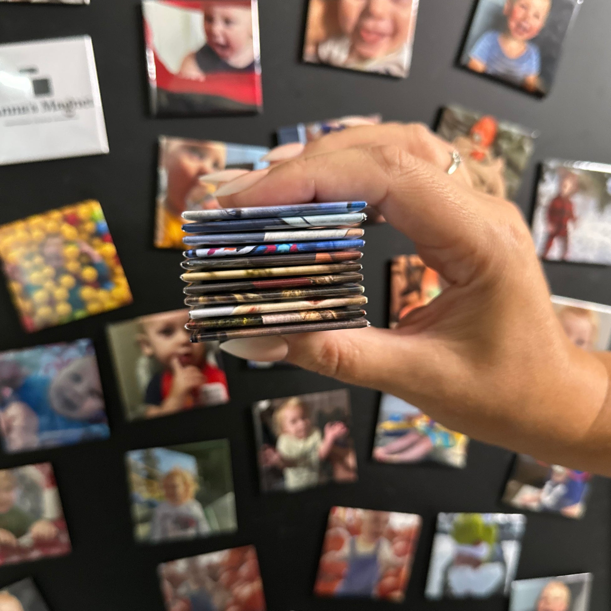 Hand holding a stack of 2x2 custom photo magnets with a background of children's photo magnets on a black board.