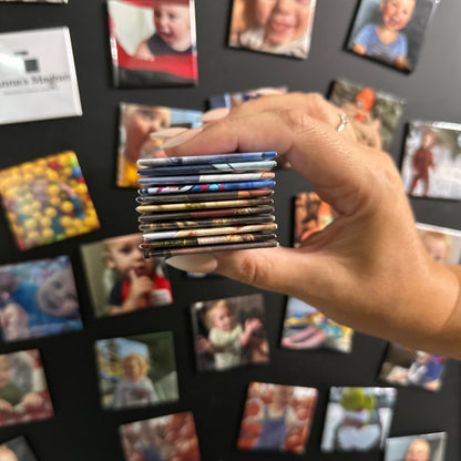 Hand holding a stack of 2x2 custom photo magnets with a background of children's photo magnets on a black board.