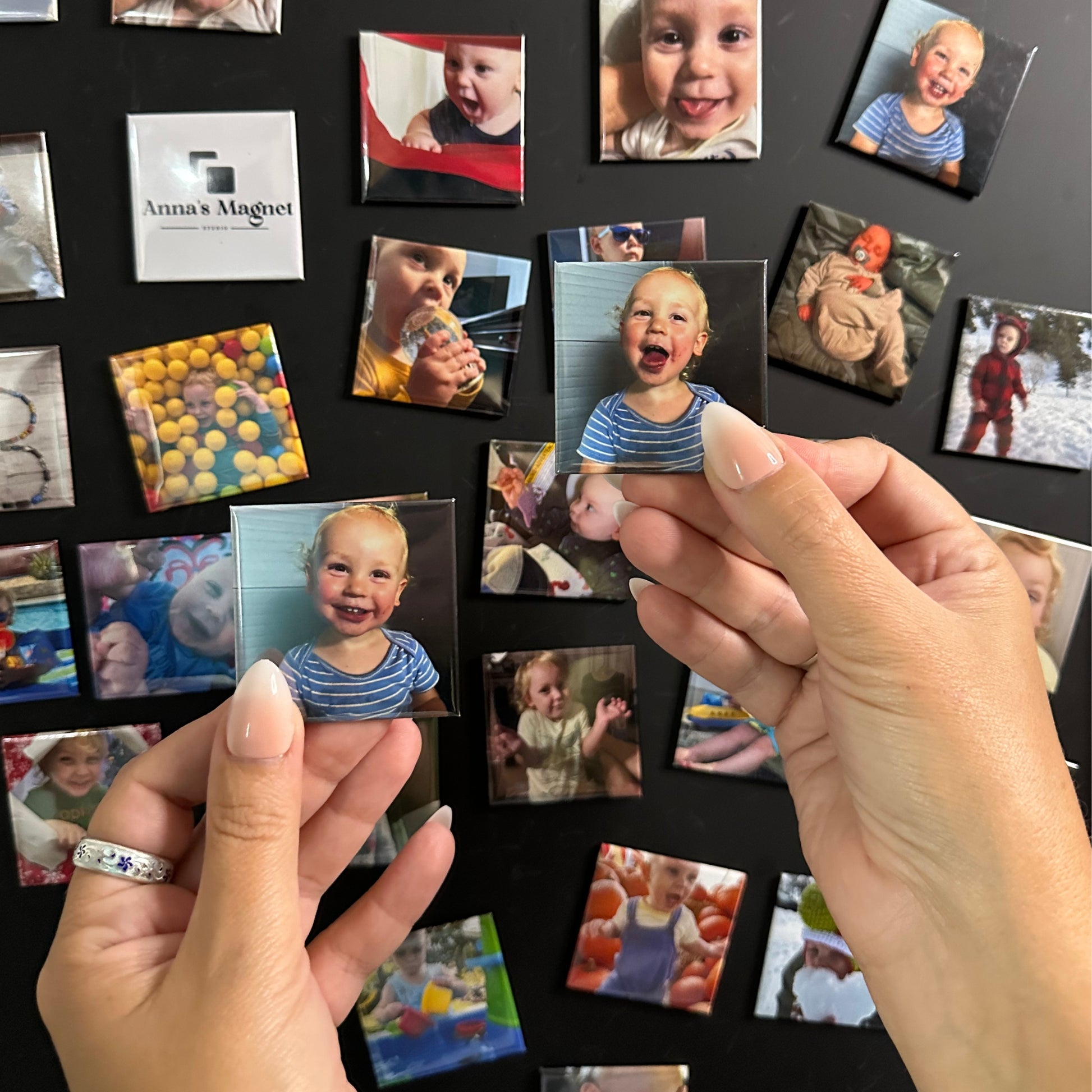 Hands holding two 2x2 custom photo magnets of a child in front of a collection of other photo magnets on a magnetic blackboard.