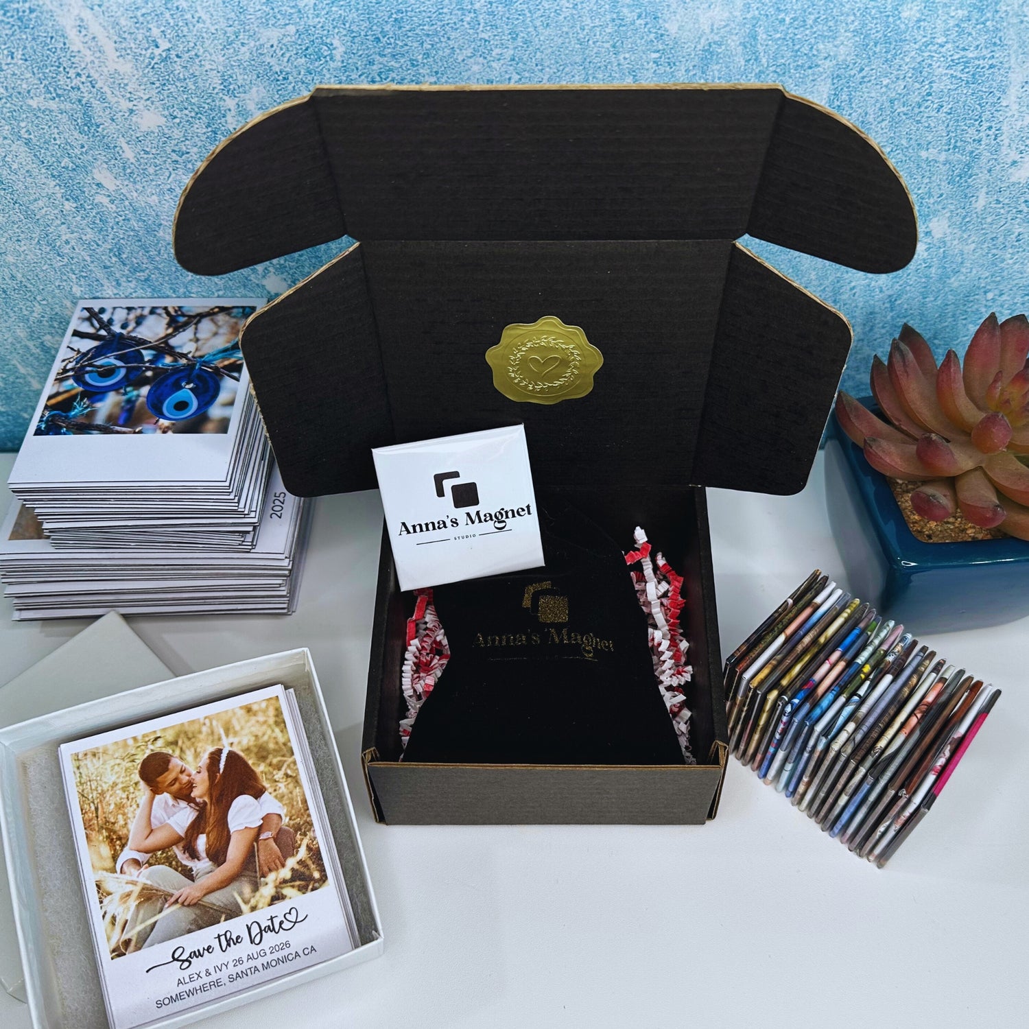 Open black box with Anna Magnet branding, surrounded by custom photo magnets and a business card on a light blue textured background.