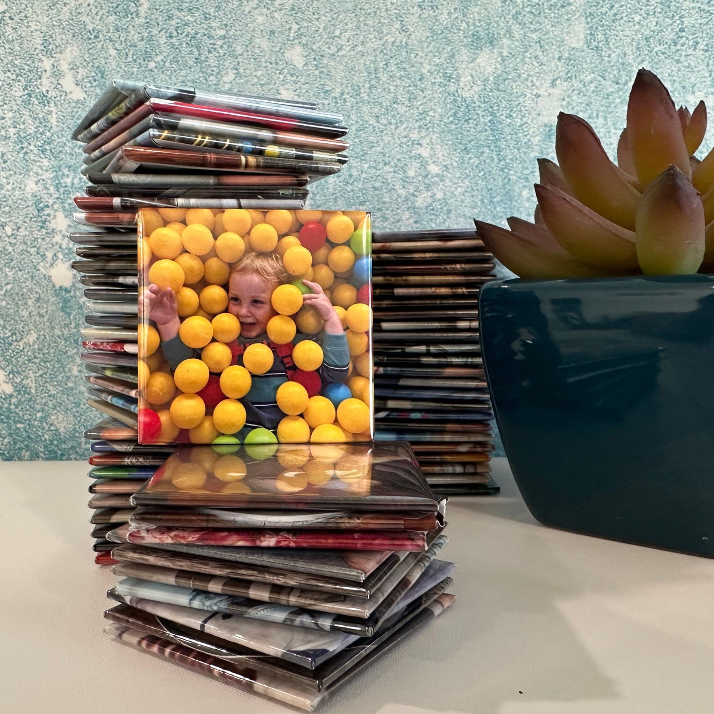 Stack of 2x2 custom photo magnets with a colorful magnet in front featuring a child among balls, placed on a surface with a plant in the background.